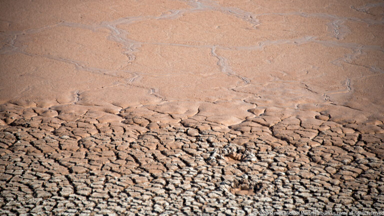 Fine polygonal cracks transition into rough, broken plates across a dry pink-tan basin floor.
