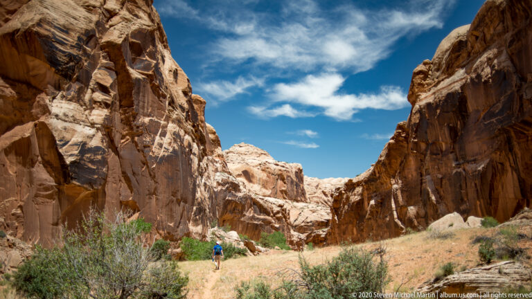 Dark vertical runoff lines mark a reddish sandstone cliff beneath pale caprock and above sparse shrubs.