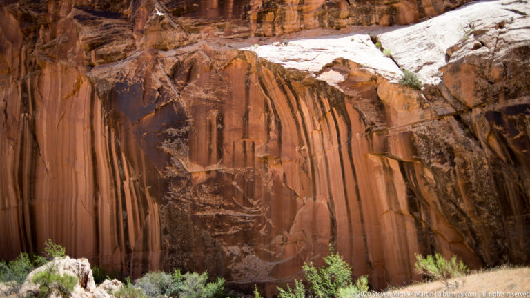 A sandy wash with shrubs leads between red sandstone walls toward a sunlit opening beneath blue sky.