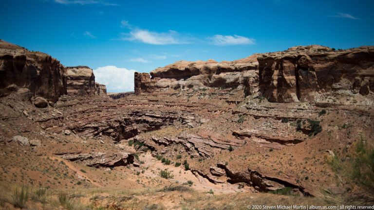 A smiling man in a pale sun hat and blue shirt poses before a softly blurred sandstone canyon wall.