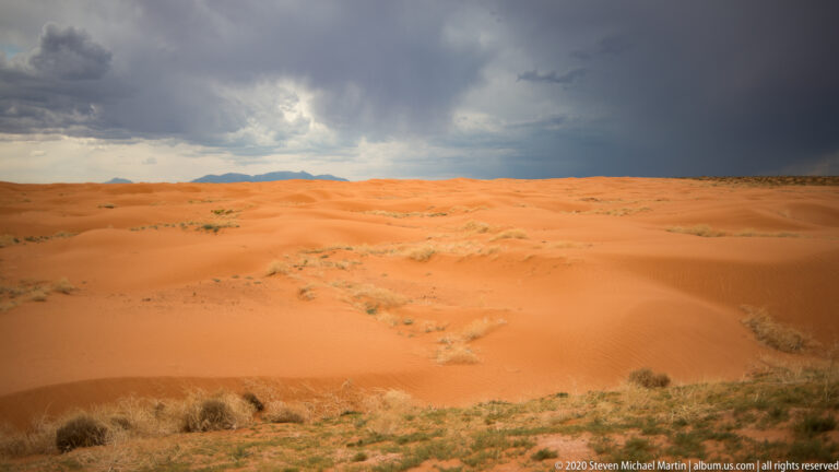 Broad orange dunes and sparse plants lie beneath a dark storm front, with distant mountains on the horizon.