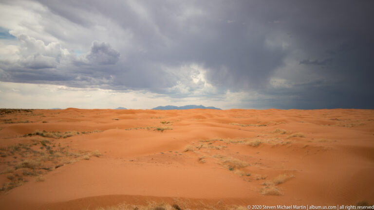 Broad orange dunes and sparse plants lie beneath a dark storm front, with distant mountains on the horizon.