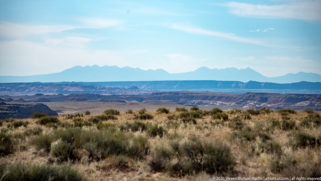 Sagebrush-covered plateau overlooks layered canyon country and a distant blue mountain range.