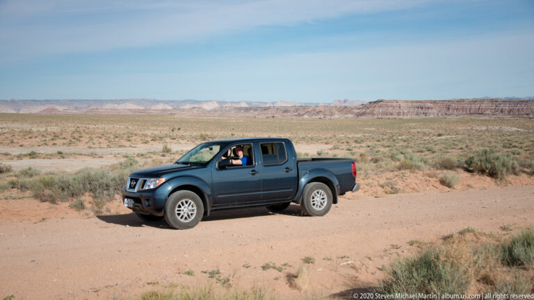 A dark pickup with a visible occupant is stopped on a sandy track across open desert scrub.