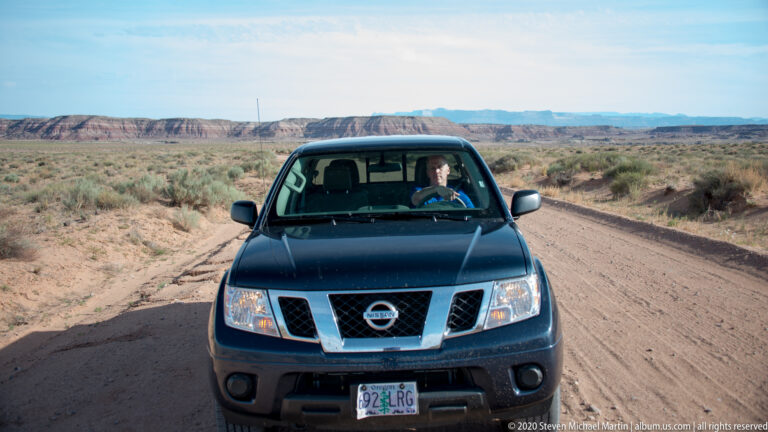 A dark pickup with a driver faces the camera on a dirt road through low scrub and open mesa country.