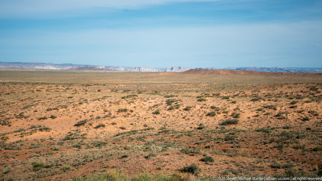 A broad scrub-covered plain extends toward pale mesas beneath a clear blue sky.