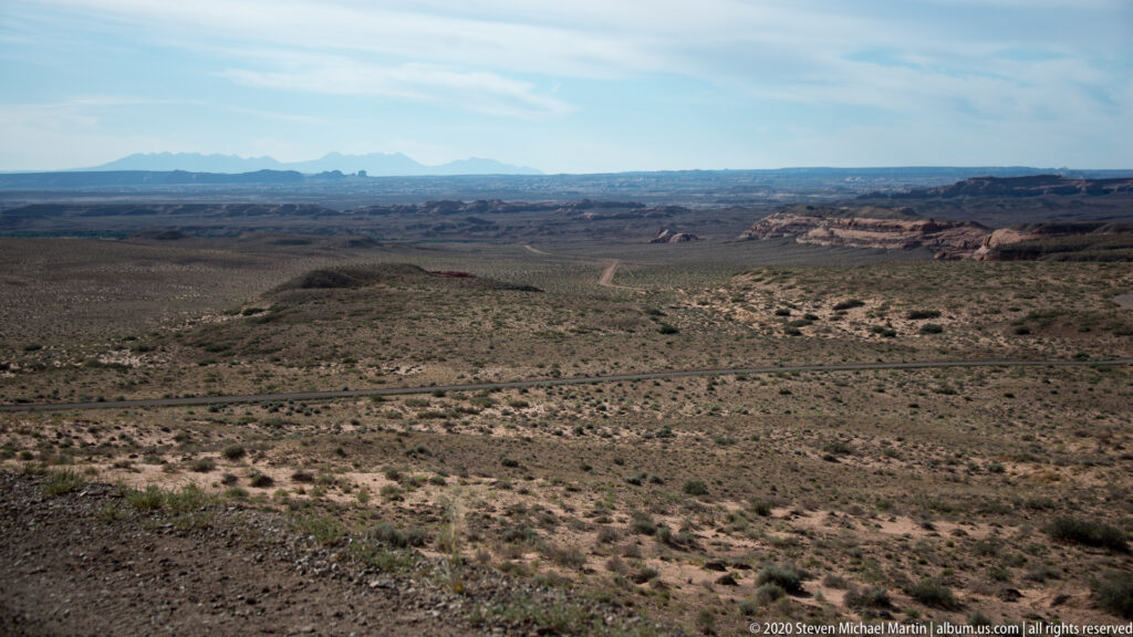 An elevated panorama crosses a canyon basin toward distant mesas and blue mountains, with a road below.