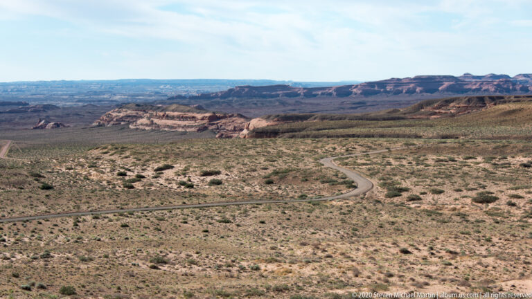 A pale road curves across scrub-covered ground toward layered red mesas on the horizon.