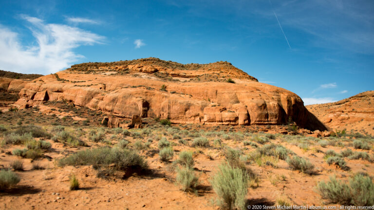 A shallow shadowed alcove interrupts an orange sandstone slope above sparse desert shrubs.