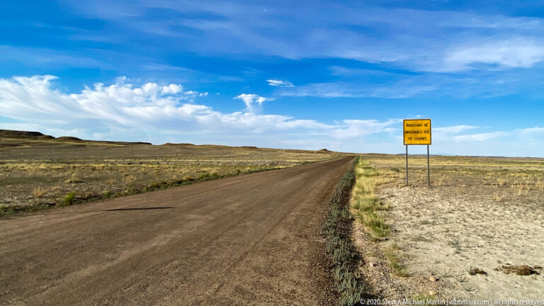 A dirt road crosses open grassland toward a low ridge, passing a yellow storm-related road warning sign.