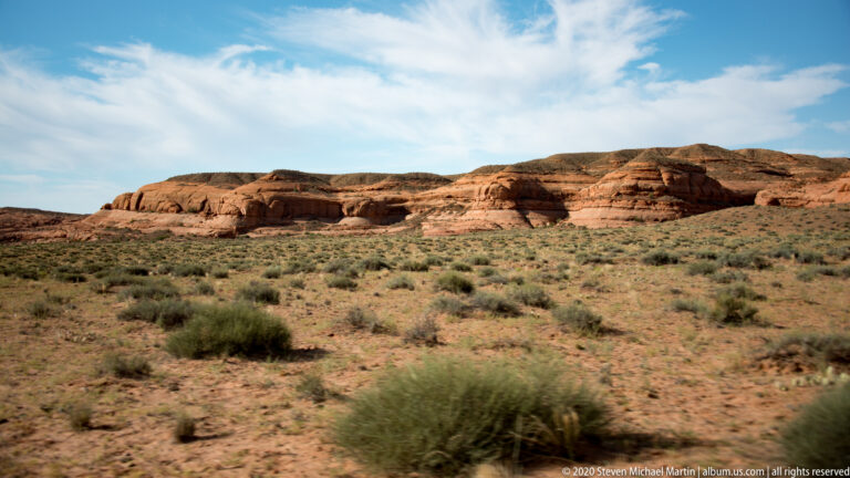 Low sandstone ridges stretch across the horizon above a wide shrub-covered desert plain beneath a blue sky.