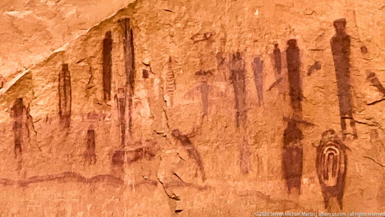 Red pictographs, including tall human-like figures and a nested oval motif, cover a sandstone wall.
