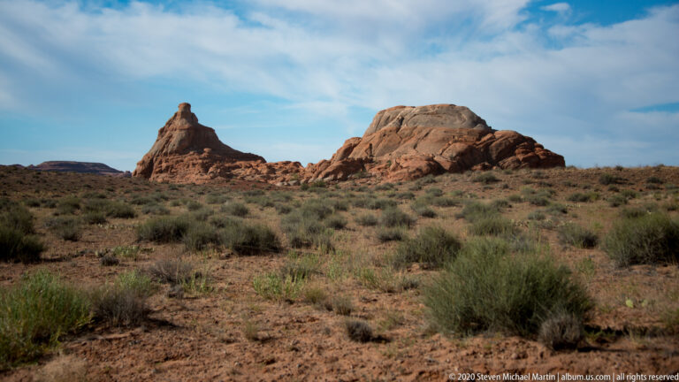 A tall sandstone butte rises from a shrub-dotted plain, with low ridges and blue sky behind it.