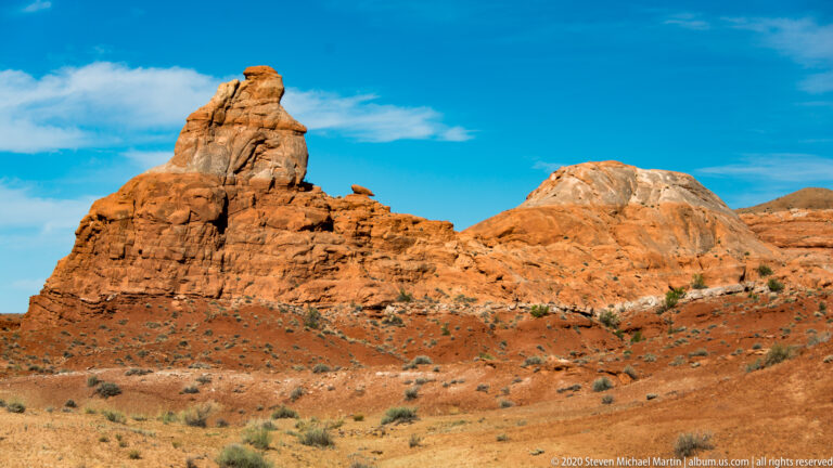 A tall red sandstone spire and a rounded rock mass rise above a shrub-dotted slickrock slope.