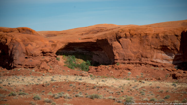 A broad low sandstone arch opens through a red rock wall above a sandy, shrub-dotted slope.