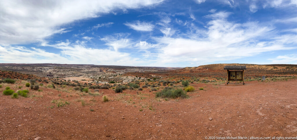 A roofed information kiosk stands on a red gravel overlook above layered canyon slopes and distant mesas.