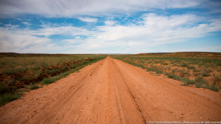 A straight red dirt road crosses open scrubland toward a distant horizon beneath broad clouds.