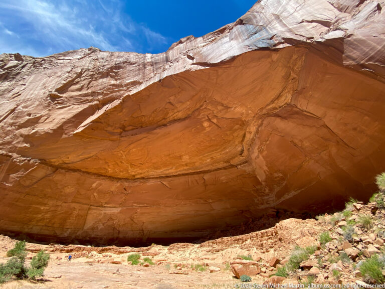 A huge layered sandstone overhang shelters a rocky floor, with a small visitor near its base for scale.