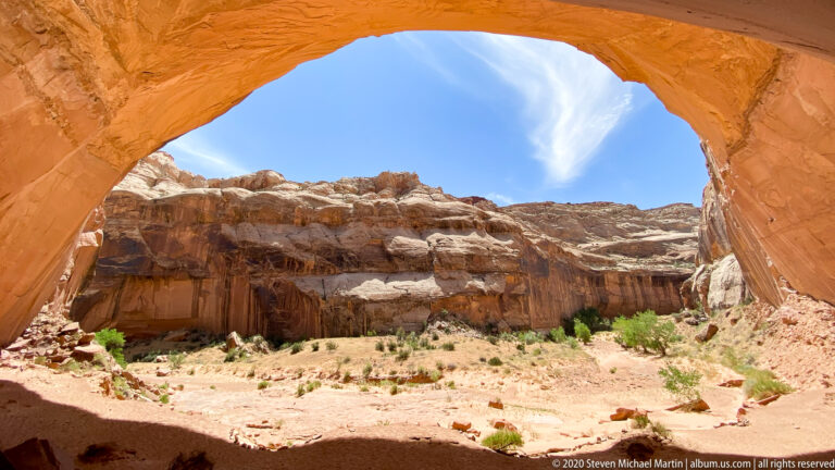 A red sandstone arch frames layered cliffs, blue sky, and sparse shrubs beyond its curved opening.