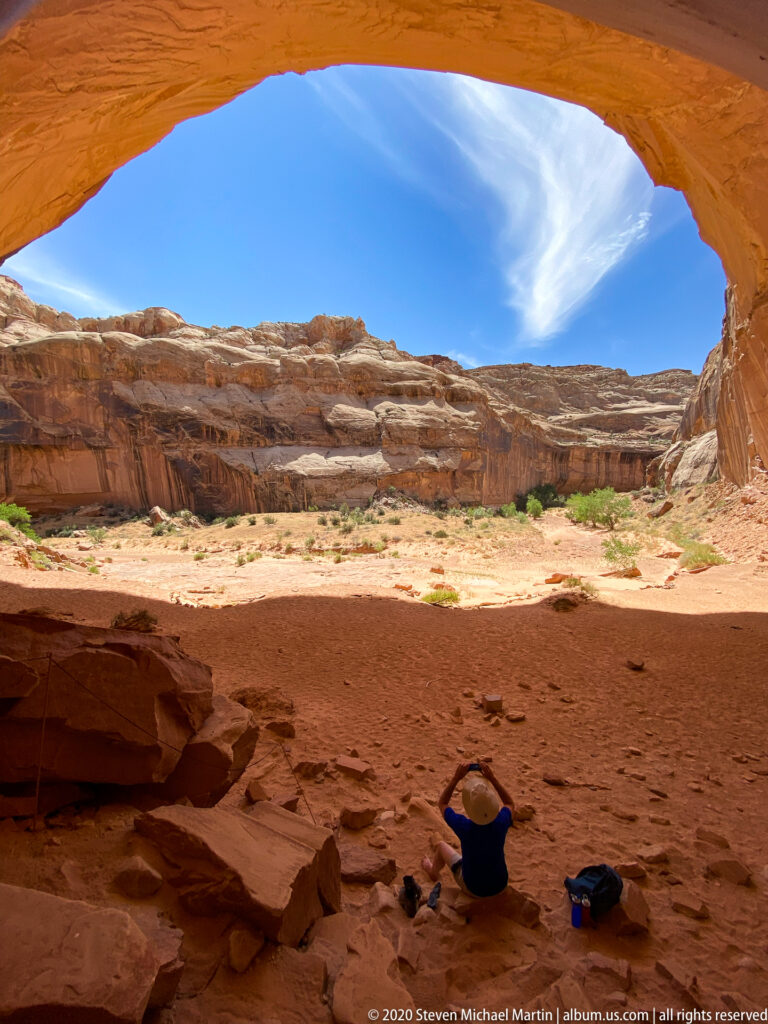 A seated visitor rests with arms raised beneath a huge sandstone overhang, with gear beside them.