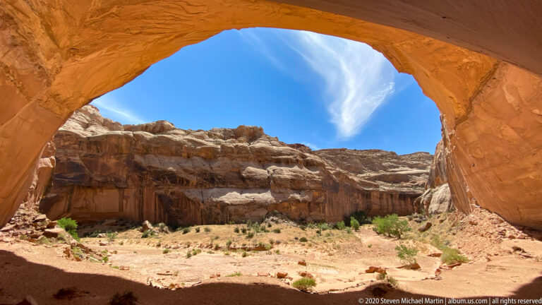 A curved sandstone arch frames a sunlit cliff and sandy canyon floor with scattered shrubs.