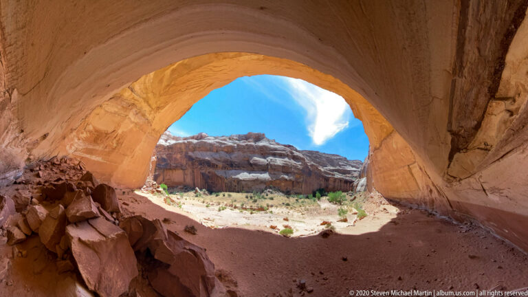 A shaded arch interior frames a sunlit sandstone wall and sandy canyon floor beyond.