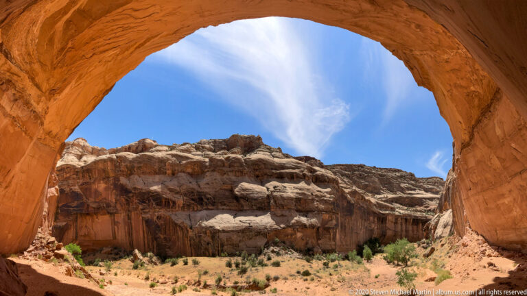 A broad orange arch curves around a view of pale cliffs, blue sky, and a dry canyon floor.
