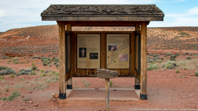 A roofed wooden kiosk with maps and interpretive panels stands in red desert scrubland.