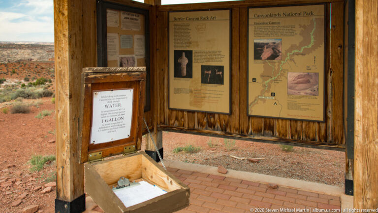Inside a wooden kiosk, rock art panels, a canyon map, water notice, and visitor register share the walls and shelf.