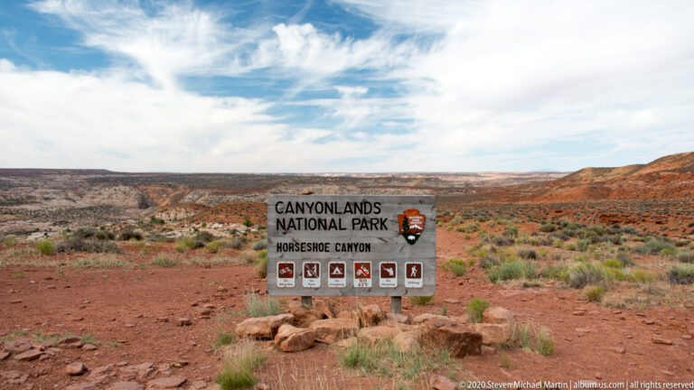 A wooden Canyonlands National Park sign names Horseshoe Canyon and displays visitor activity symbols against desert mesas.