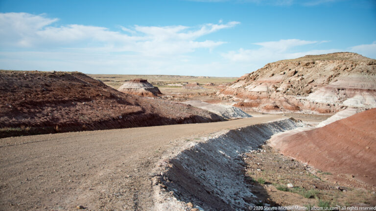 A pale dirt road bends through layered white, gray, and red badlands beneath a clear sky.