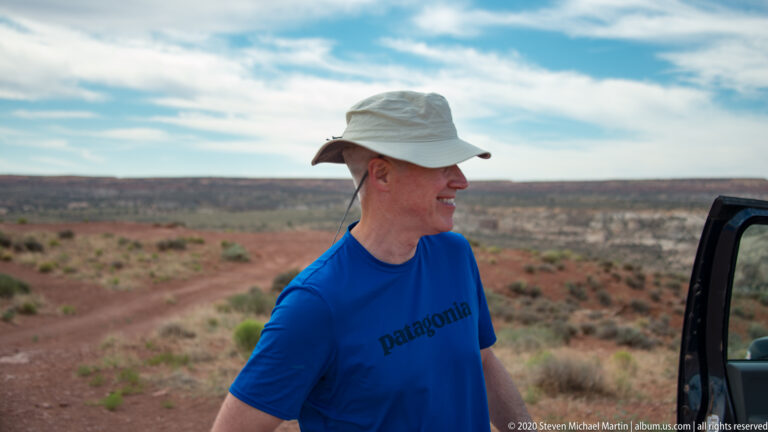 A man in a pale sun hat and blue shirt stands beside an open vehicle door in the desert.