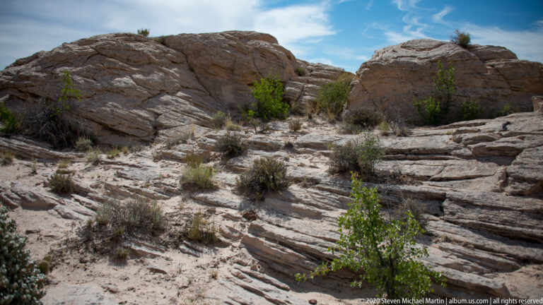 Pale layered sandstone slabs descend among small shrubs toward rounded boulders under a blue sky.
