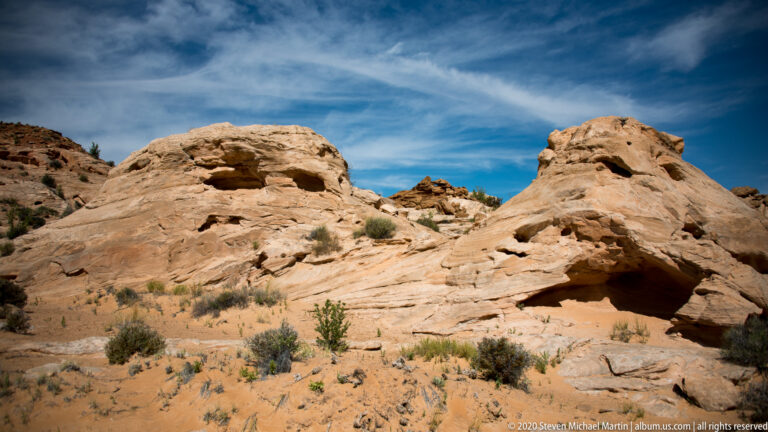 Rounded orange sandstone formations with shallow cavities and overhangs rise above a sandy wash.