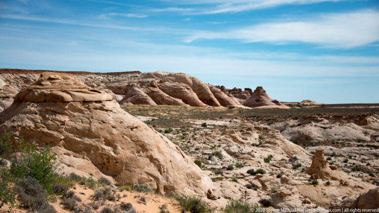 A pale foreground shelf overlooks striped sandstone ridges and a broad, sparsely vegetated basin.