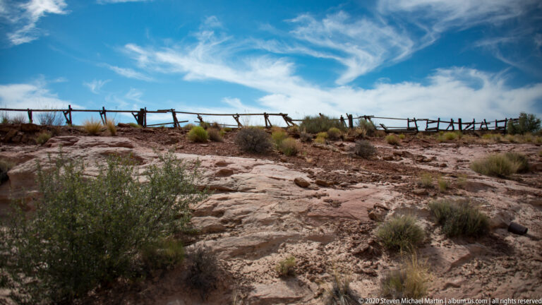 A weathered split-rail fence crosses the ridge above a rocky path and low desert shrubs.