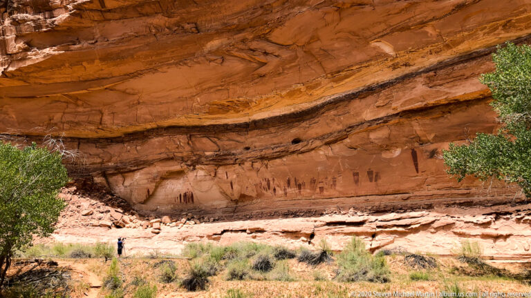 A visitor stands below a long sandstone panel of dark figures and marks beneath a projecting ledge.