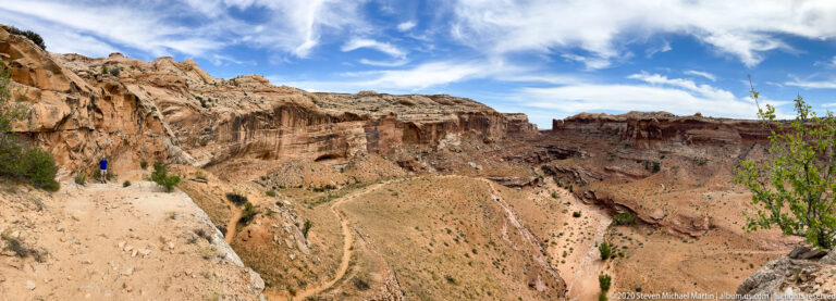 A lone hiker stands beside a tall sandstone wall above a narrow, winding trail into the canyon.