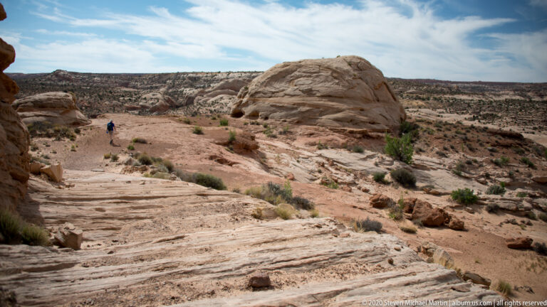 A hiker crosses a rocky sandy trail toward a rounded sandstone dome beside a distant canyon rim.
