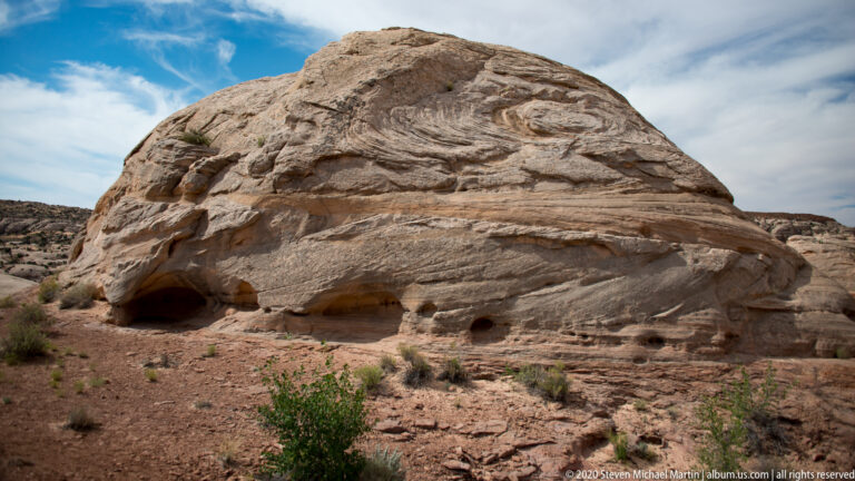 A pale rounded sandstone dome with shallow dark cavities rises above sandy ground and sparse shrubs.