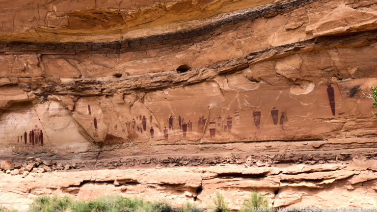 Dark elongated figures and smaller marks cross a red sandstone wall beneath a horizontal rock ledge.