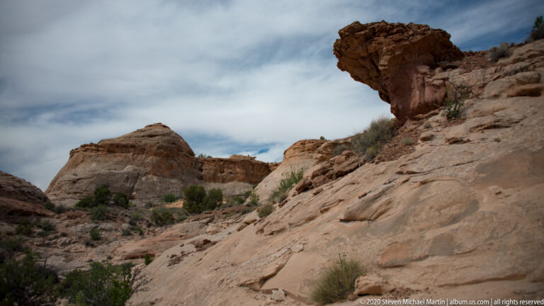 A heavy sandstone ledge projects above a sloping rock face and narrow shrub-lined canyon passage.