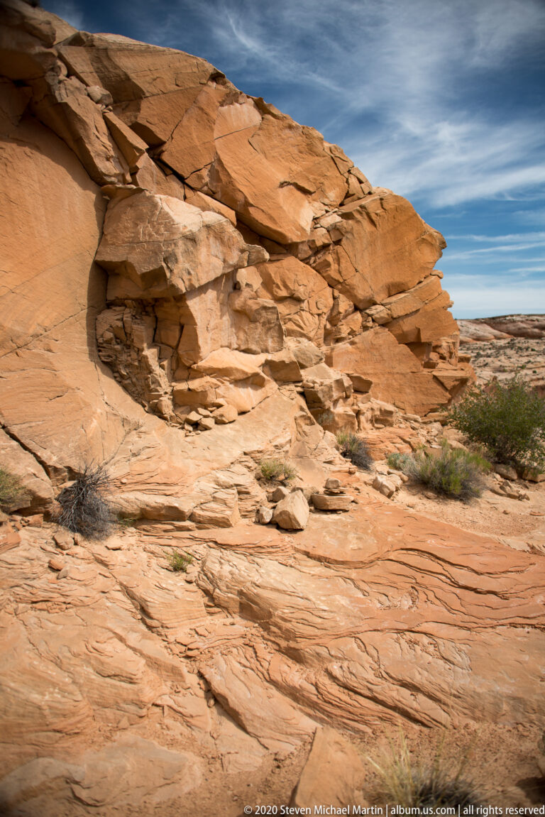 An orange sandstone ledge projects over broken slabs, with layered rock and a flat rim beyond.