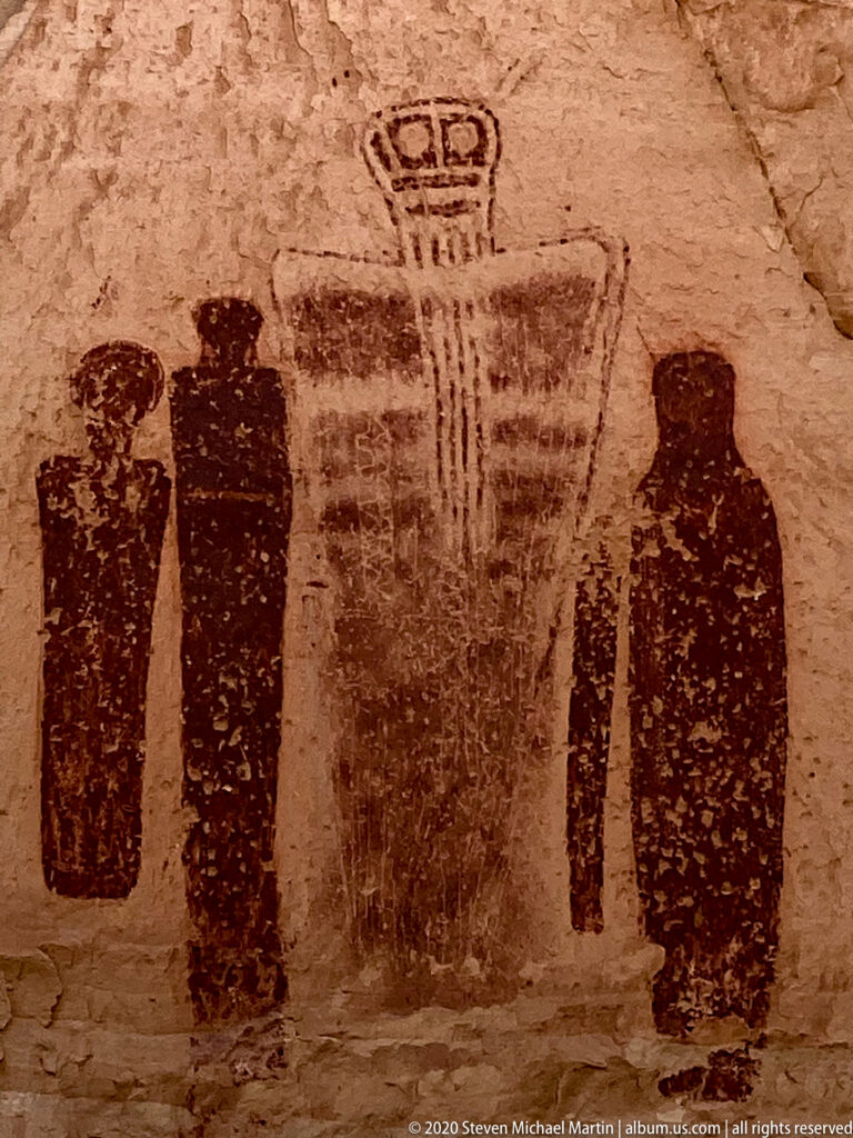 Dark elongated figures surround a broad central painted form topped by a looped motif on sandstone.