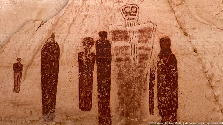 Tall dark figures flank a larger central figure with a looped element on a cracked sandstone wall.