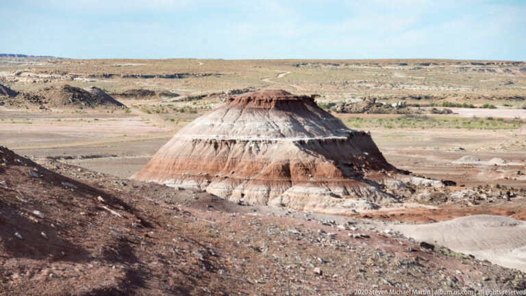 A flat-topped, banded butte rises above an open basin of sparse scrub and eroded sediment.