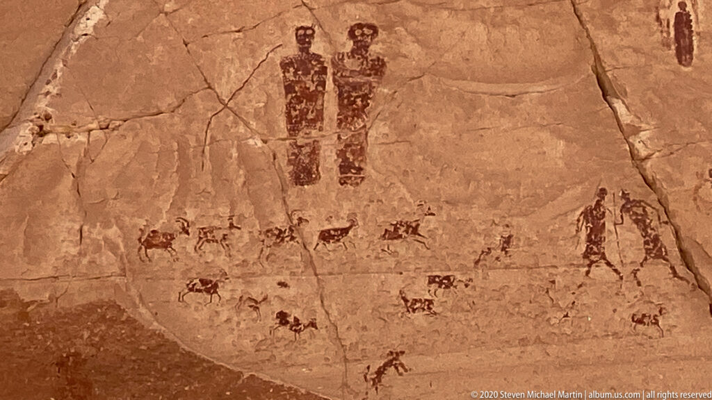 Two tall figures appear above scattered animal forms, with a larger outline figure at right on sandstone.