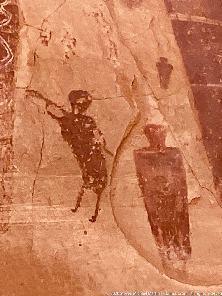A dark broad-bodied figure with extended limbs stands beside a blocklike painted form on sandstone.