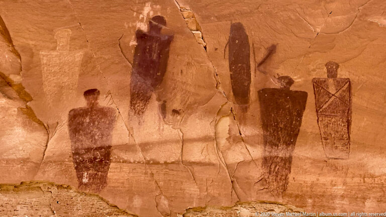 Tall dark figures, including a crosshatched form, stand across a cracked sandstone wall.
