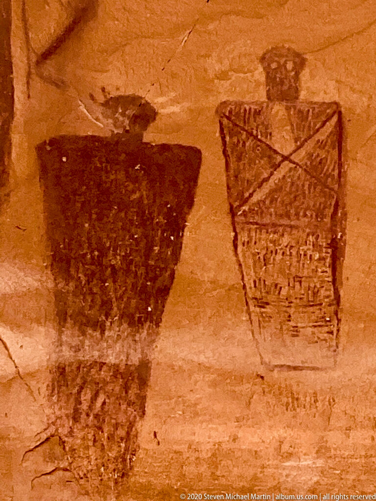 Two large dark painted forms sit on sandstone; one is solid and broad, the other patterned with crossed lines.